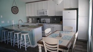 A compact kitchen with a central island, bar stools, a dining table with chairs, and a clock on the wall. - Sunset Square O - Second Row complex in Garden City Beach, SC (Garden City)