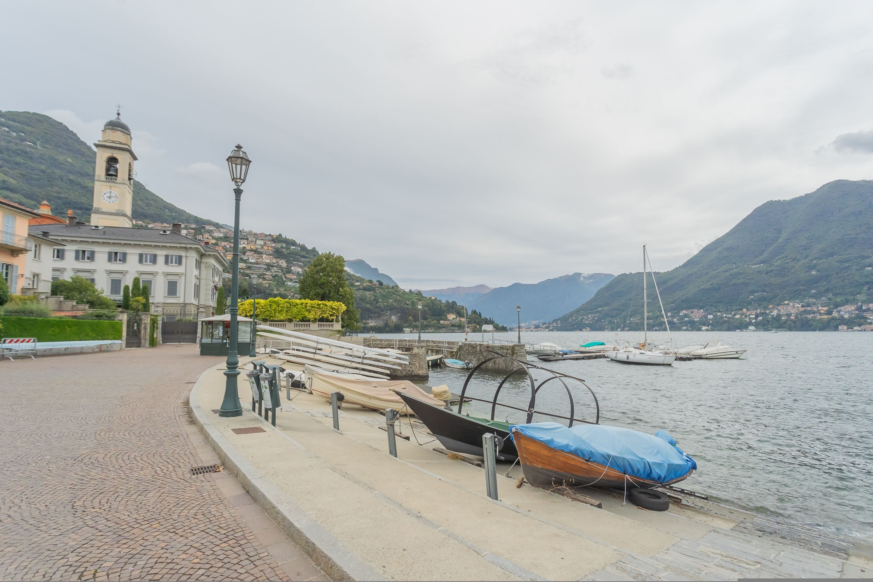 A waterfront promenade with boats docked, a clock tower, and a mountainous backdrop.