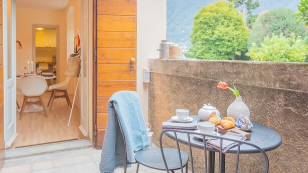 A balcony with a table set for tea, a chair with a towel, and a view of greenery and mountains. - La Piazza di Cernobbio (Cernobbio)