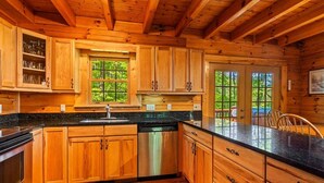 A wooden cabin-style kitchen with a dishwasher, granite countertops, and a window overlooking greenery. - Smokey Mountain Retreat (Waynesville)