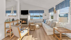 Amplia sala de estar con chimenea, un conjunto de sofás, televisor y ventanas grandes con vista a un cuerpo de agua.