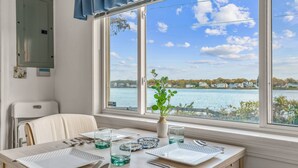 Mesa de comedor dispuesta con platos, cubiertos y vasos, ubicada junto a una ventana con vista a un lago y edificios.