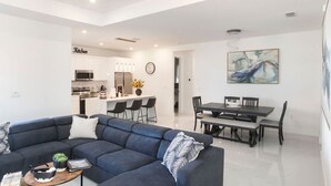 Ein modernes Wohnzimmer mit einem dunkelblauen Modulsofa, einem Essbereich mit einem dunklen Holztisch und Stühlen sowie einer Küche mit weißen Schränken und Edelstahlgeräten.