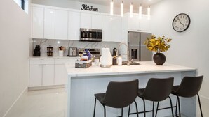 Eine moderne Küche mit weißen Schränken, einer zentralen Kochinsel inklusive Barhocker, einer Mikrowelle, einem Kühlschrank und einer Wanduhr.