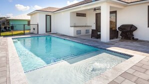 Ein rechteckiger Pool mit Fliesenrand, eine überdachte Terrasse mit Grill und ein weißes Gebäude mit flachem Dach.