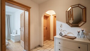 Un couloir avec une porte en bois, un banc blanc orné d’un coussin à motif de cerf, une commode blanche avec des tiroirs, un miroir et un vase avec des plantes.