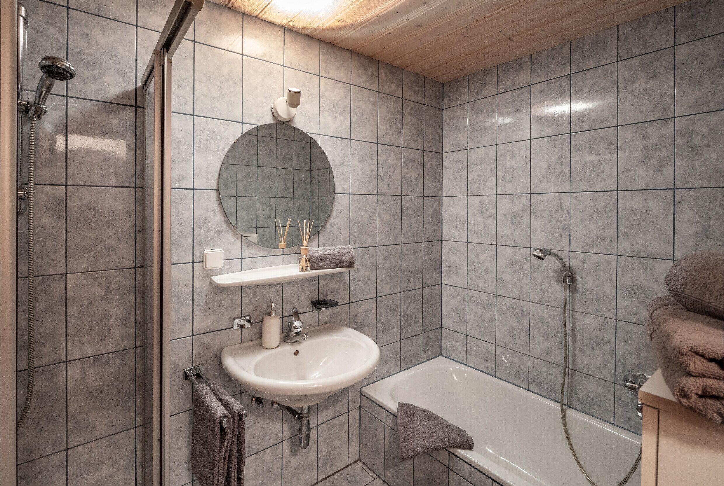 A modern bathroom with a shower, sink, and bathtub, featuring grey tiles and a wooden ceiling.