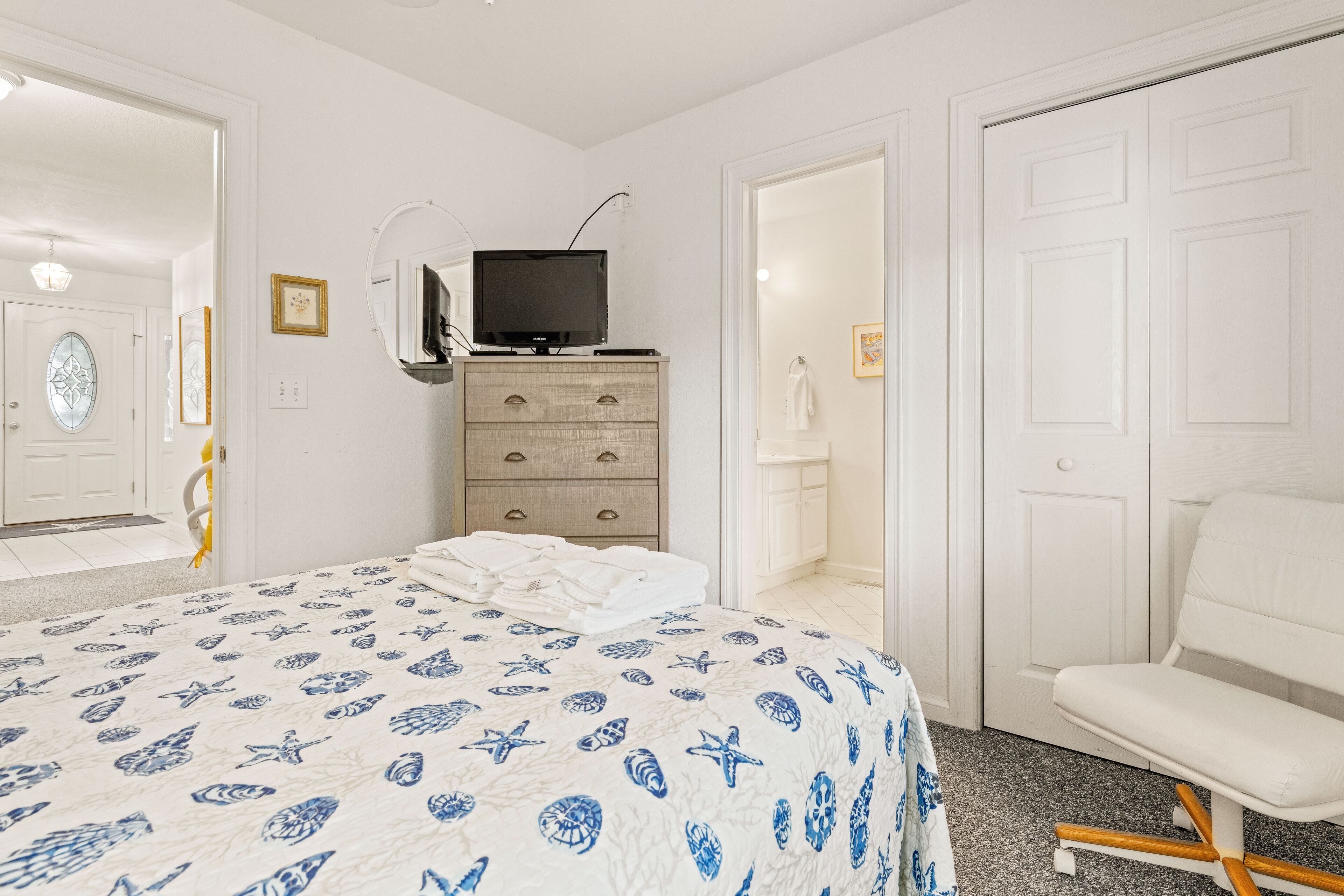 Una camera da letto con un letto, una televisione su un comò, una sedia bianca e una porta con uno specchio.