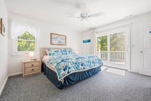 Une chambre à coucher comprenant un lit, une table de chevet, un ventilateur de plafond, une fenêtre avec des rideaux, une porte et une vue sur l’extérieur.