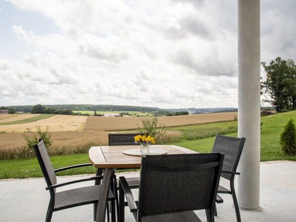 An outdoor patio with a table and chairs, a vase with flowers, and a view of fields and a cloudy sky.