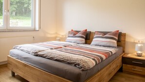 Un dormitorio con una cama de madera, una ventana con vistas a la vegetación, una mesita de noche con una lámpara y otra mesita de noche.
