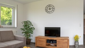 Una sala de estar con un sofá, una mesa de centro con fruta, un televisor sobre un mueble de madera, una planta y un reloj en la pared.