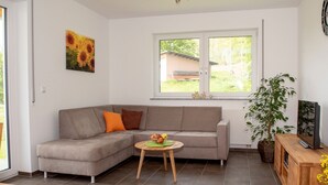 Una sala de estar moderna con un sofá seccional gris, una mesita redonda con fruta, una planta en maceta y un televisor sobre un mueble de madera.