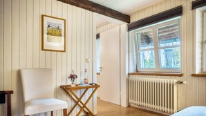 A room with a wooden floor, a white chair, a small wooden table with a bottle and flowers, a radiator, and a window with a view of greenery. - Holiday Apartment 'Alte Polsterei' with Private Terrace, Shared Garden and Wi-Fi (Nienstädt)