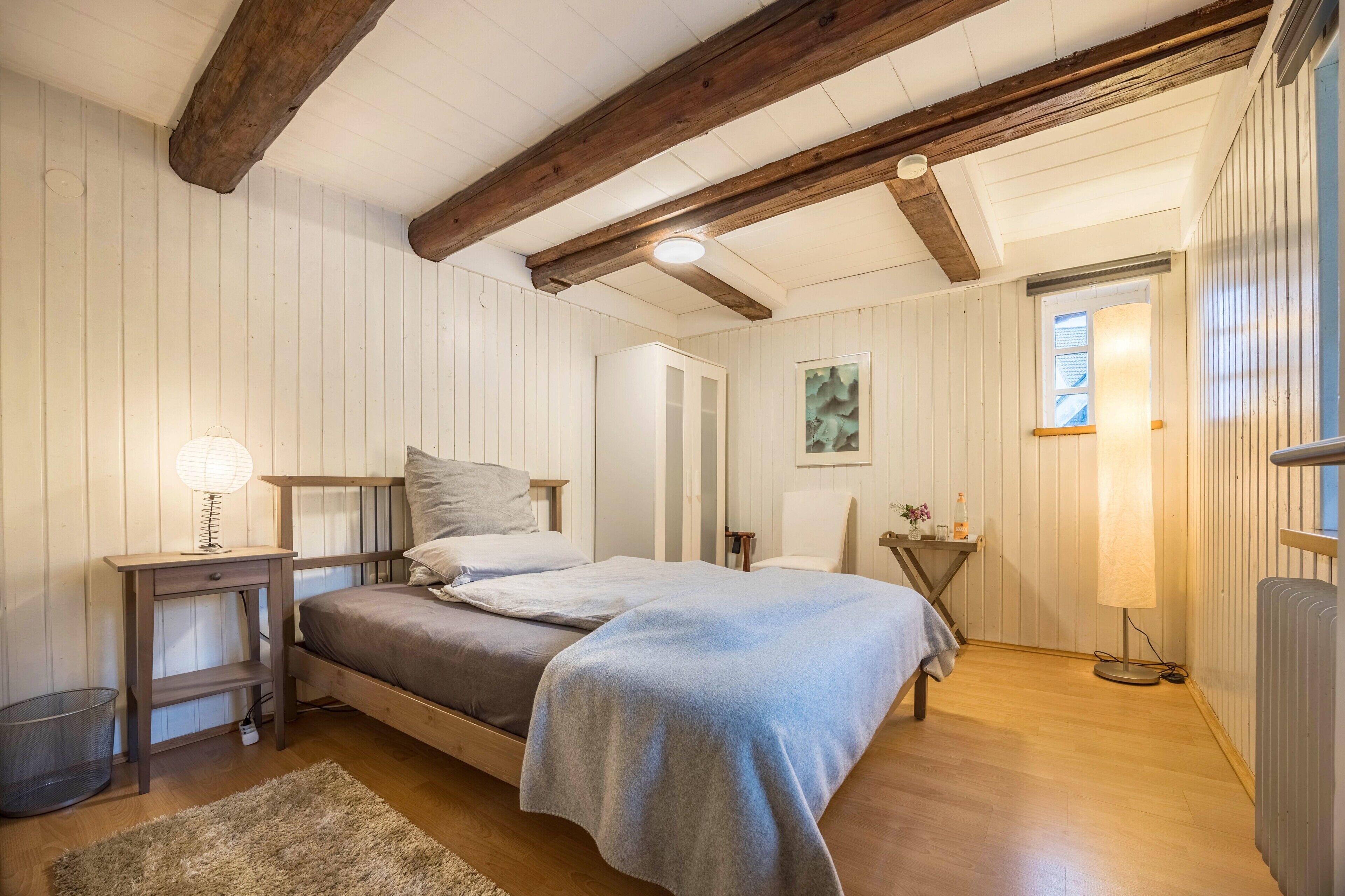Un dormitorio con cama, mesita de noche, lámpara, radiador y vigas de madera en el techo.