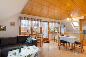 A cozy living room with a sofa, dining table, and chairs, wooden flooring, and large windows.