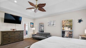A bedroom with a large bed, a ceiling fan, a TV, a dresser, a gray sofa, and a window with blinds.