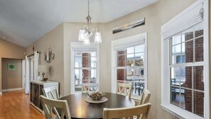A dining area with a round table, wooden chairs, and a chandelier. There are large windows with brick walls and a view of the outdoors.
