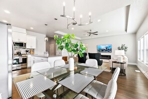 A modern kitchen with stainless steel appliances, a dining area with a glass table and striped placemats, and a living room with a TV and a ceiling fan.