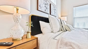 Una cama bien hecha con ropa blanca, una lámpara de noche, un control remoto y un pequeño florero con una planta sobre una mesita de noche de madera.