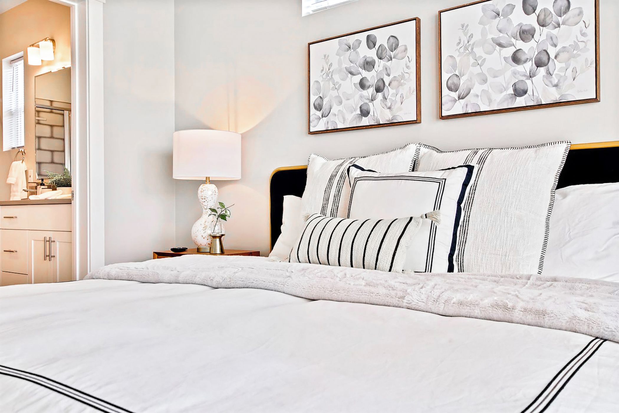 Un lit bien fait, avec une couette blanche et des oreillers à rayures, une table de chevet avec une lampe, et deux œuvres d’art encadrées accrochées au mur au-dessus du lit.