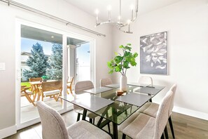 A modern dining area with a glass table, cushioned chairs, a potted plant, and a chandelier.