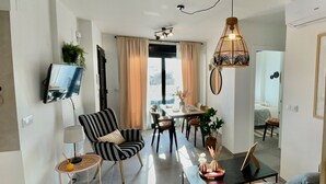 A modern living room with a dining area, a TV mounted on the wall, and a large window with curtains. - Anacasa Macomer Oliva Playa AP803 (Oliva)