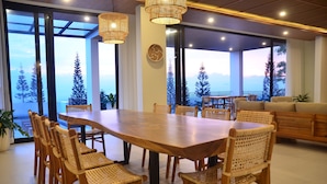 Un vaste espace dédié à la salle à manger, avec une grande table en bois et des chaises en osier, de grandes fenêtres offrant une vue sur les arbres, et un balcon doté d’une balustrade.