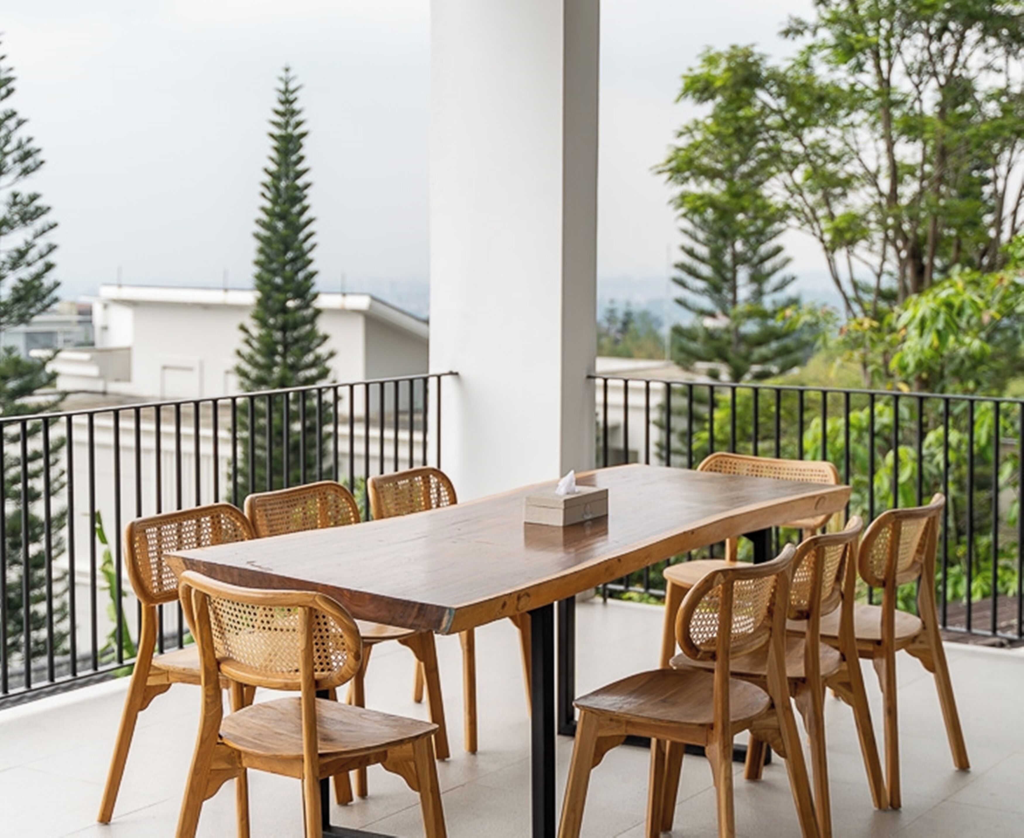 A wooden table with six chairs on a balcony with a black railing, overlooking a residential area with trees and buildings.