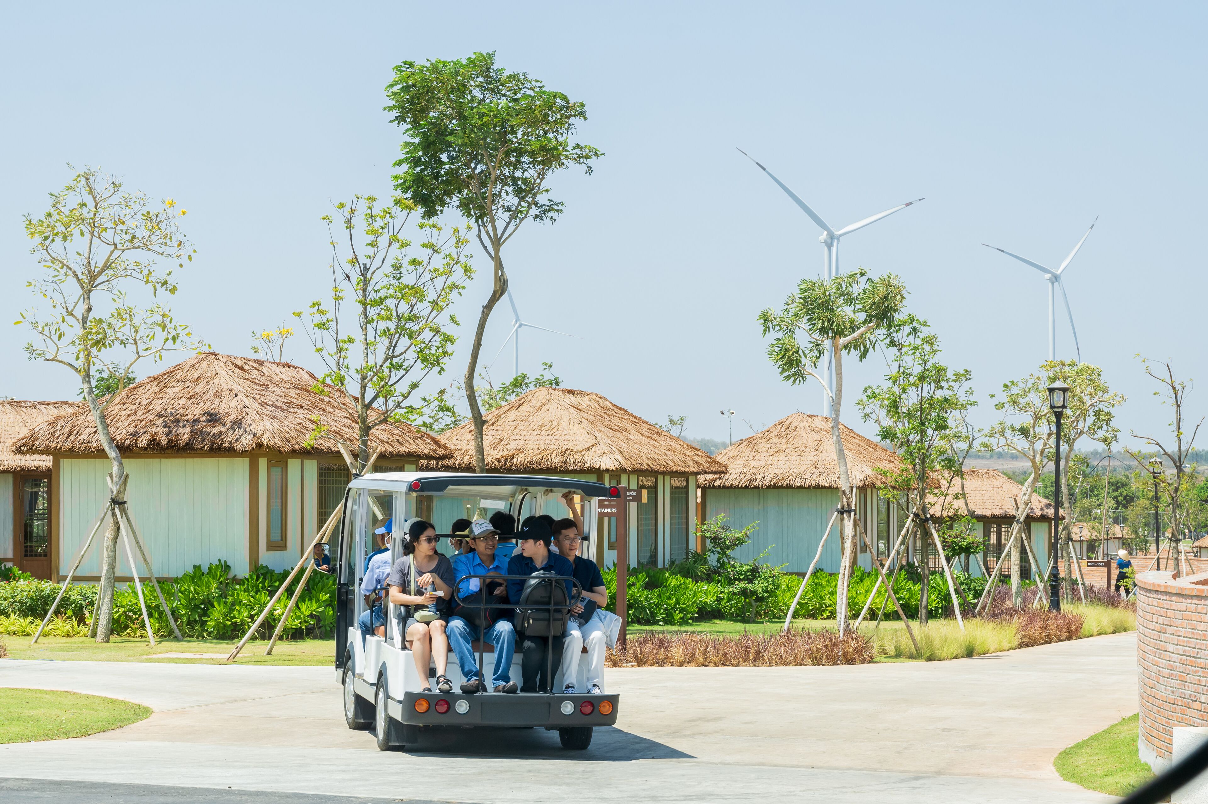 A tour vehicle with passengers, surrounded by thatched-roof buildings and wind turbines.