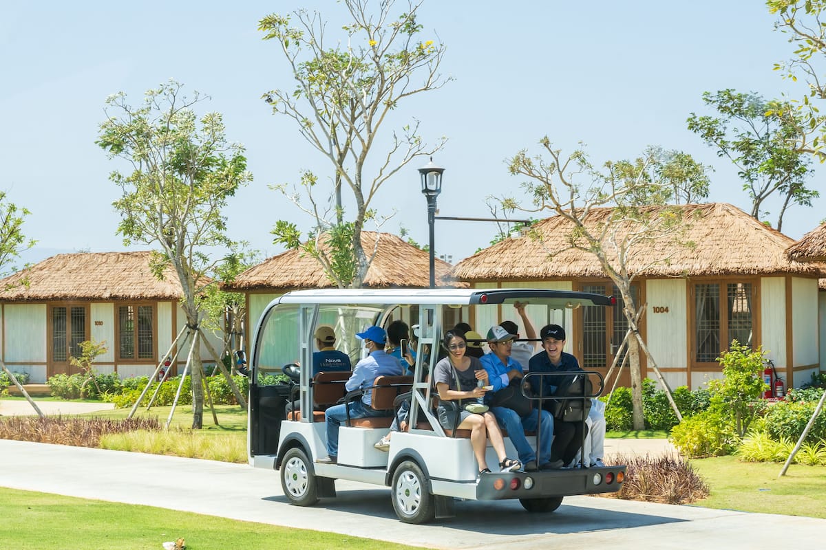 A group of people on a golf cart tour through a resort area with thatched-roof buildings and well-maintained gardens.