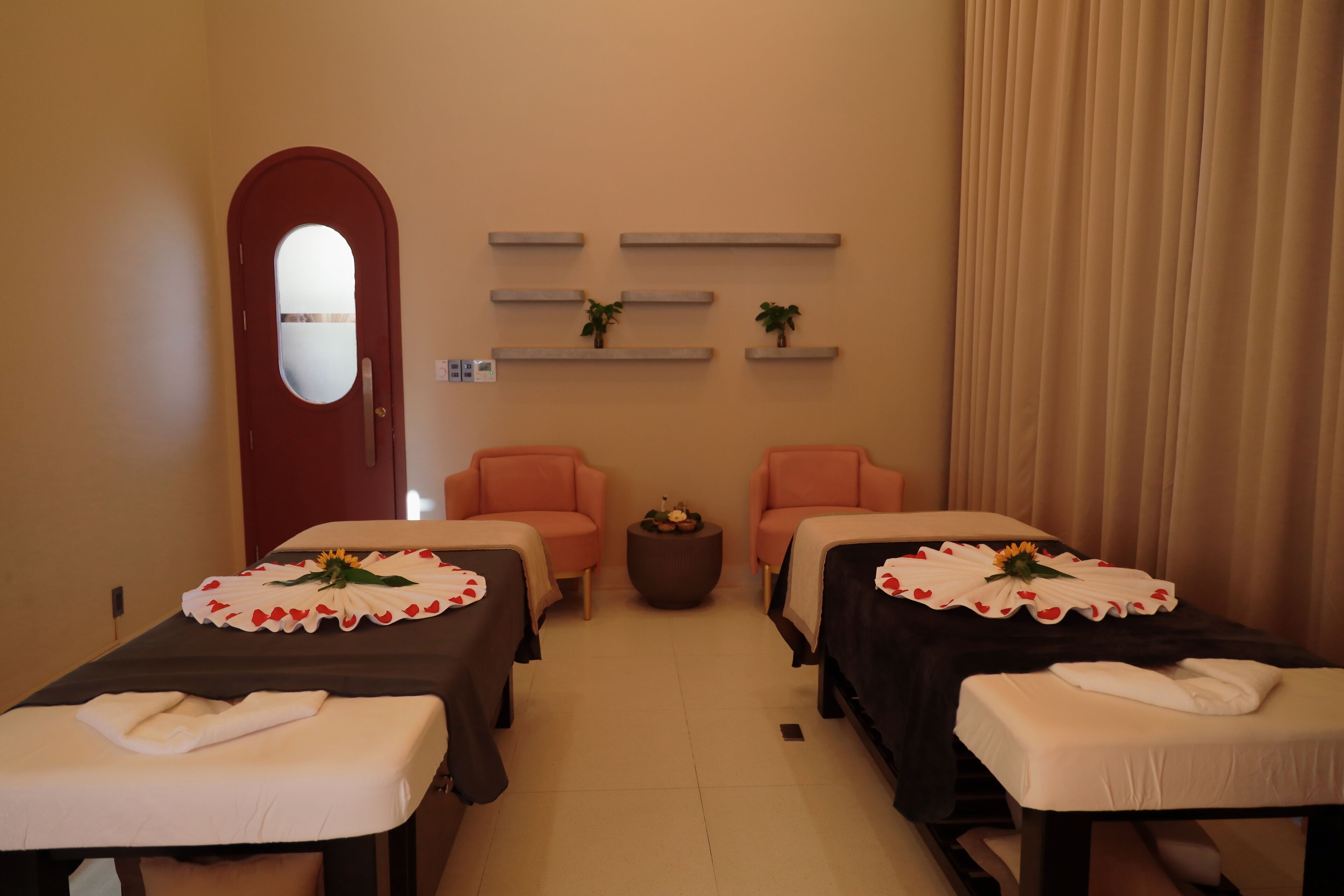 A room with two massage tables, each with a towel and a flower arrangement. There are two chairs and a small round table with a plant. Shelves with decorative items are on the wall.
