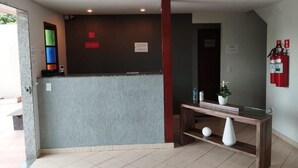 A reception area with a grey reception desk, a wooden console table with a vase and a plant, and a fire extinguisher mounted on the wall. - Suite Pousada Paraío (Guarapari)
