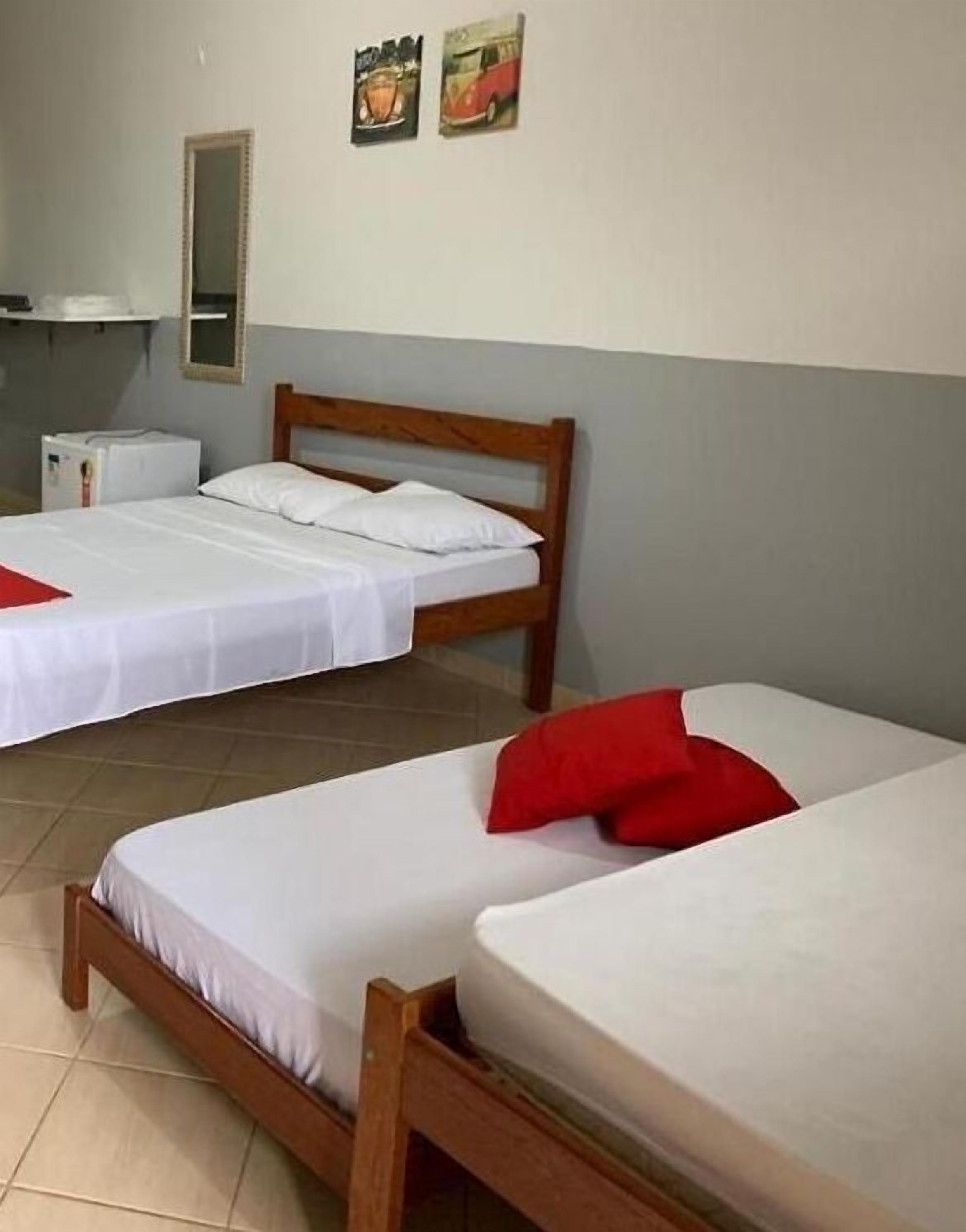 A room with two wooden beds, white bedding, and a red pillow on one bed. There is a small white cabinet and a mirror on the wall.