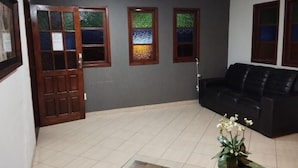 A room with a black leather sofa, a wooden coffee table, and a potted plant. There are three windows with stained glass and a door with a wooden frame. - Suite Pousada Paraío (Guarapari)