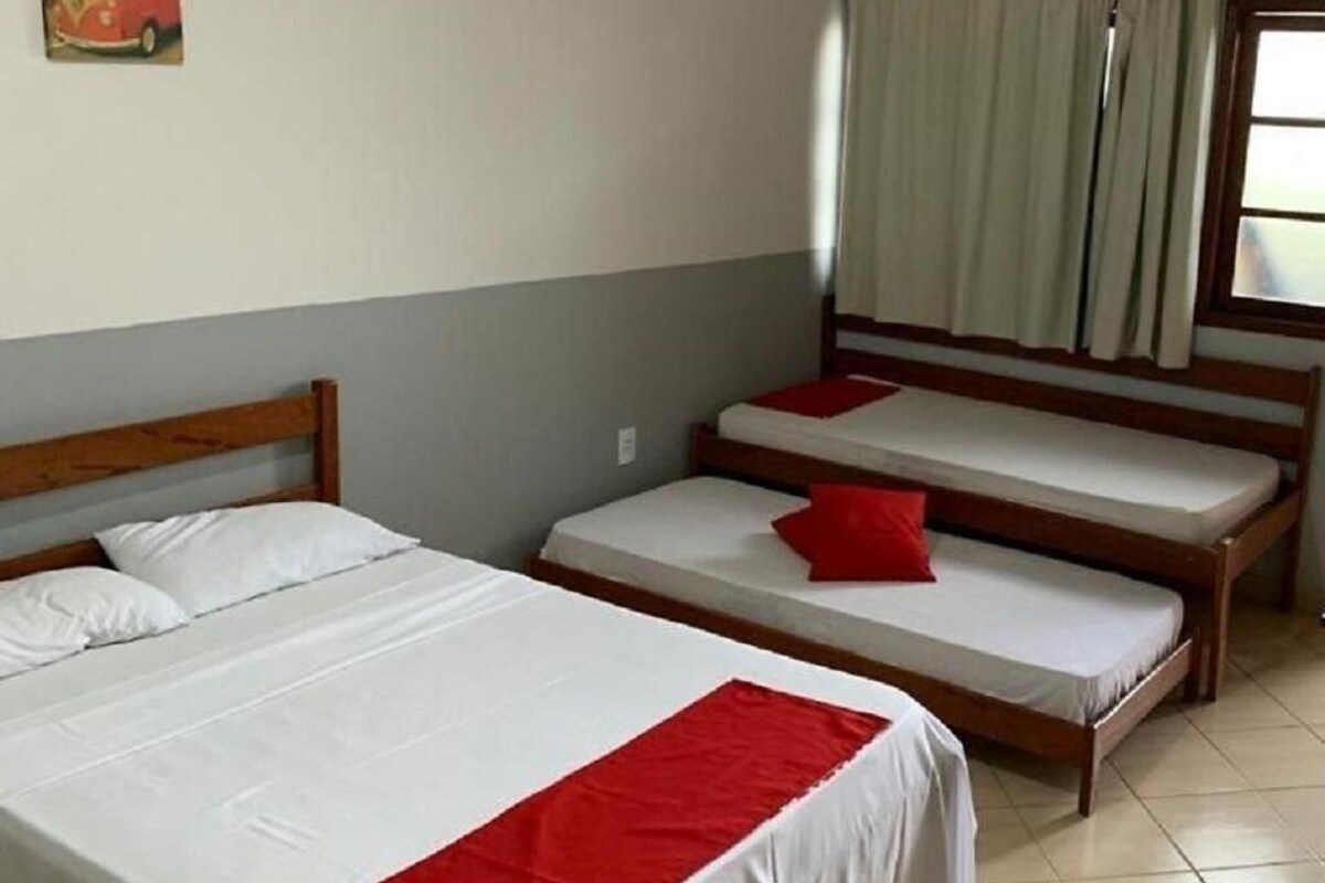 A room with two single beds and a larger bed, all with white bedding and red accents. There is a window with curtains, a wall-mounted picture, and a tiled floor.