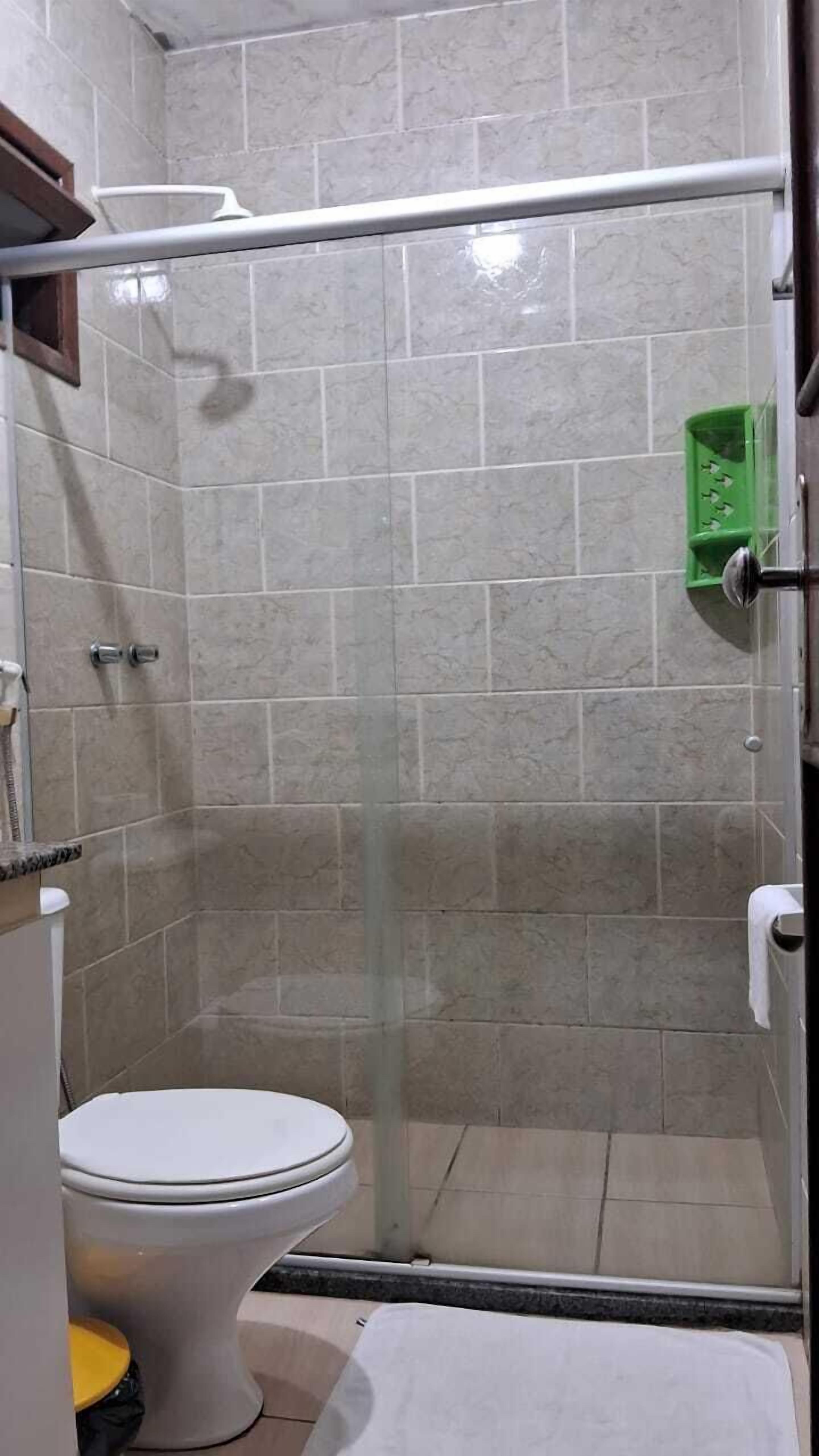 Um banheiro com chuveiro em box de vidro, vaso sanitário e uma lixeira verde.