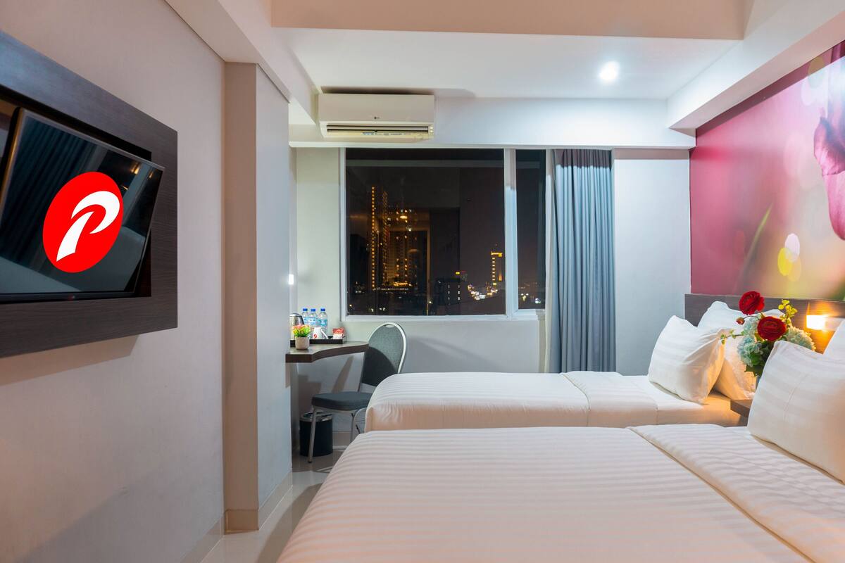 Ein Hotelzimmer mit zwei Betten, einem Schreibtisch, einem Sessel, einem Fernseher und einem großen Fenster mit Blick auf die Stadt.