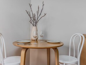 Een ronde houten tafel met twee witte stoelen, een vaas met takken en twee wijnglazen.