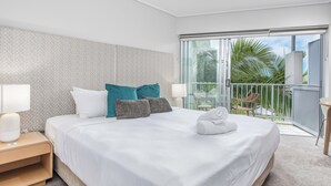 A hotel room with a large bed, a nightstand, a lamp, and a view of the balcony with palm trees. - The Sand Resort Kingscliff - Studio Apt (Tweed)