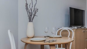 A dining area with a round table, white chairs, a vase with branches, a television, and a wooden cabinet. - The Sand Resort Kingscliff - Studio Apt (Tweed)