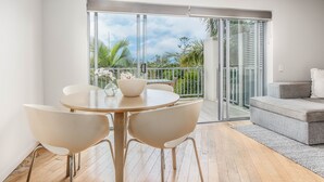 Una sala de estar moderna con una mesa redonda, sillas blancas, un sofá gris y un gran ventanal corredero que da a un balcón con vistas a palmeras y al cielo.