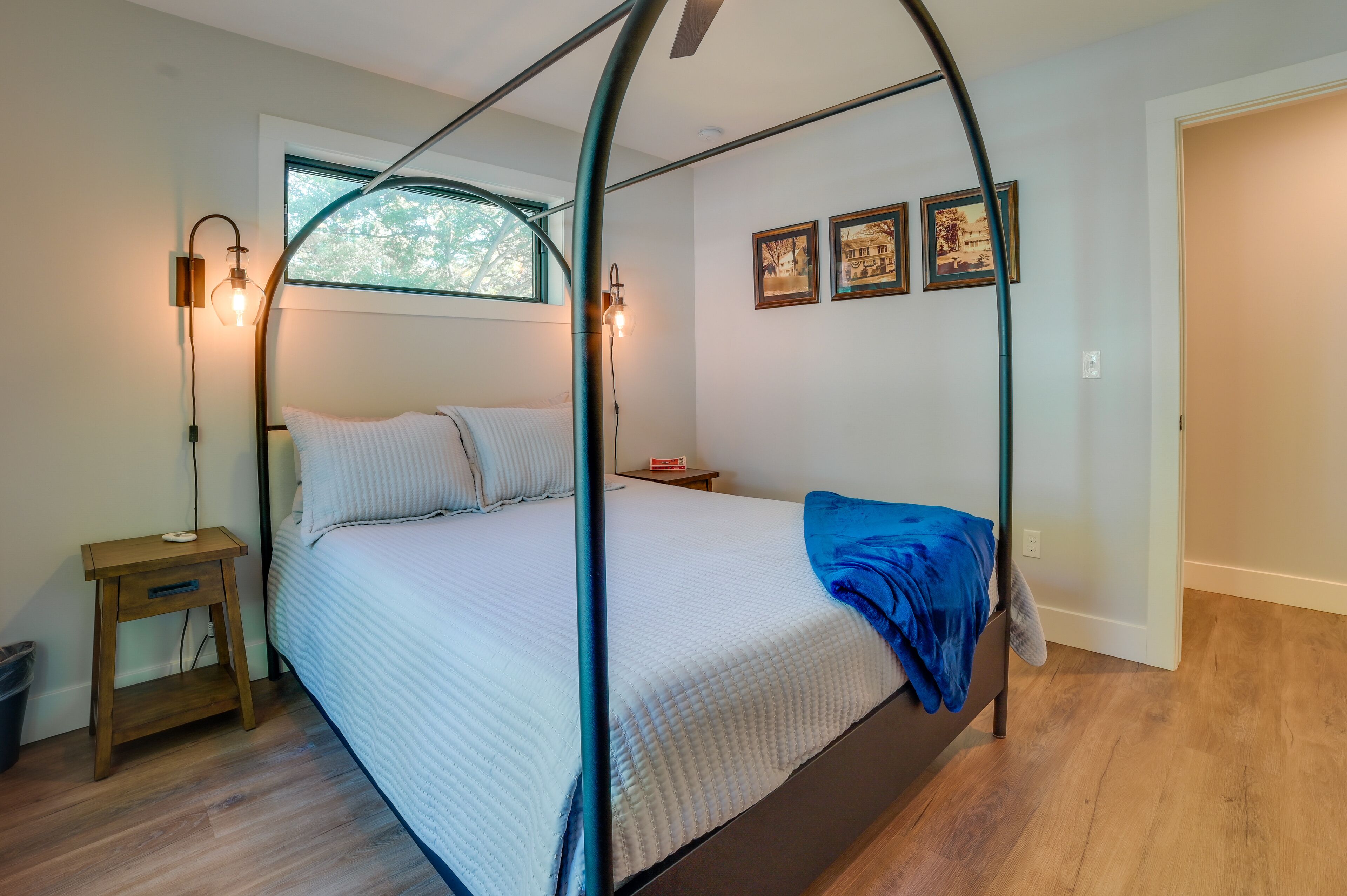 A bedroom with a four-poster bed, a nightstand, a lamp, and a window with a view of trees.