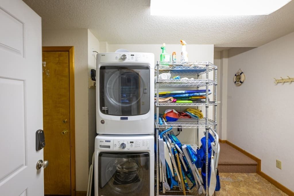 Une buanderie équipée de machines à laver et de sèche-linge superposés, d’étagères contenant des produits de nettoyage et d’une porte donnant sur une autre pièce.