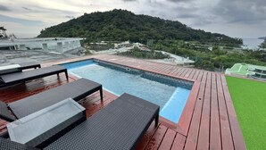 Une piscine entourée de chaises longues sur une terrasse en bois, avec une colline verdoyante en arrière-plan et une vue sur une ville côtière.