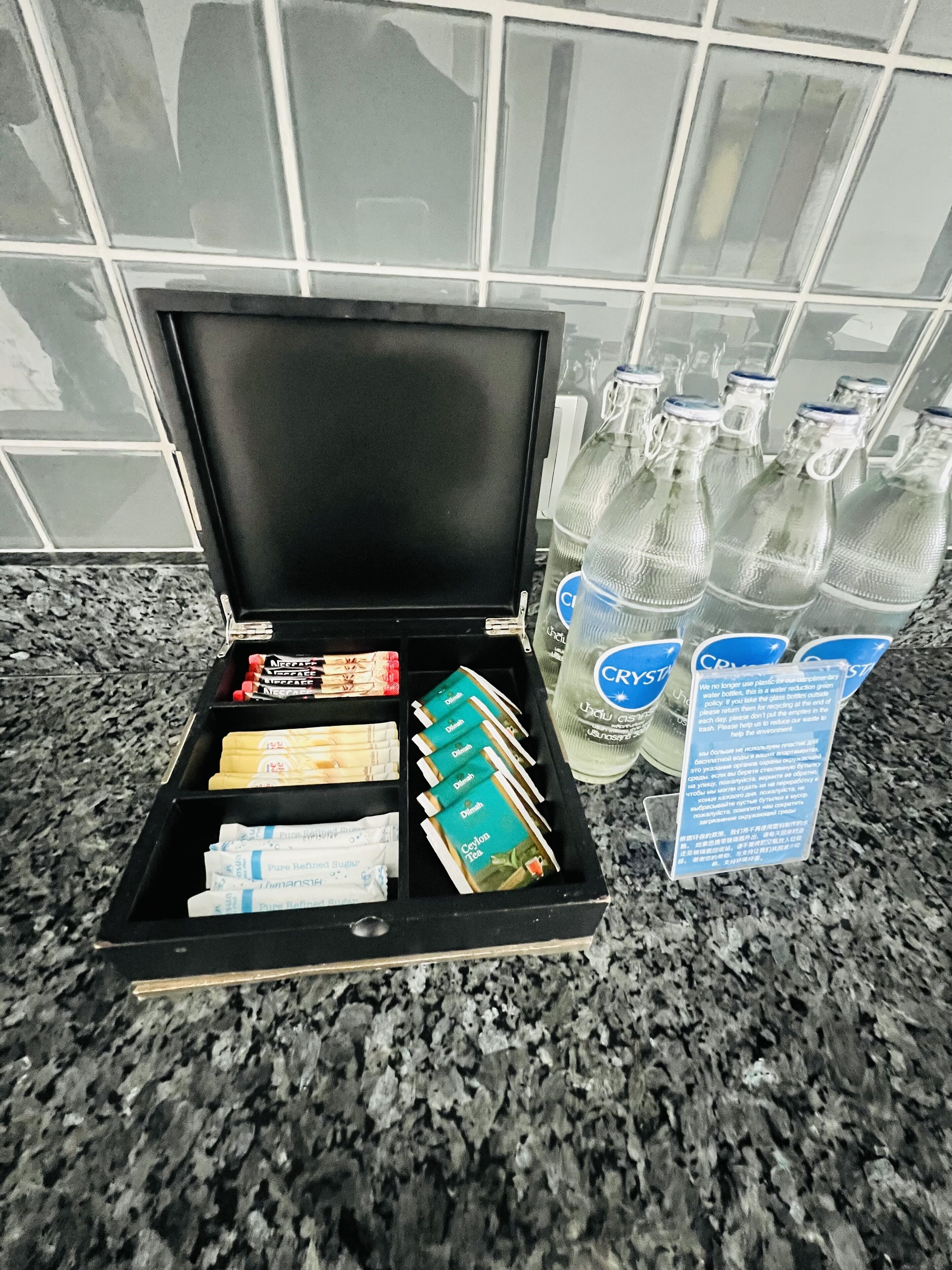 Un plateau noir contenant divers articles, notamment des bouteilles d’eau, des sachets de thé et un mot posé sur un comptoir en granit.