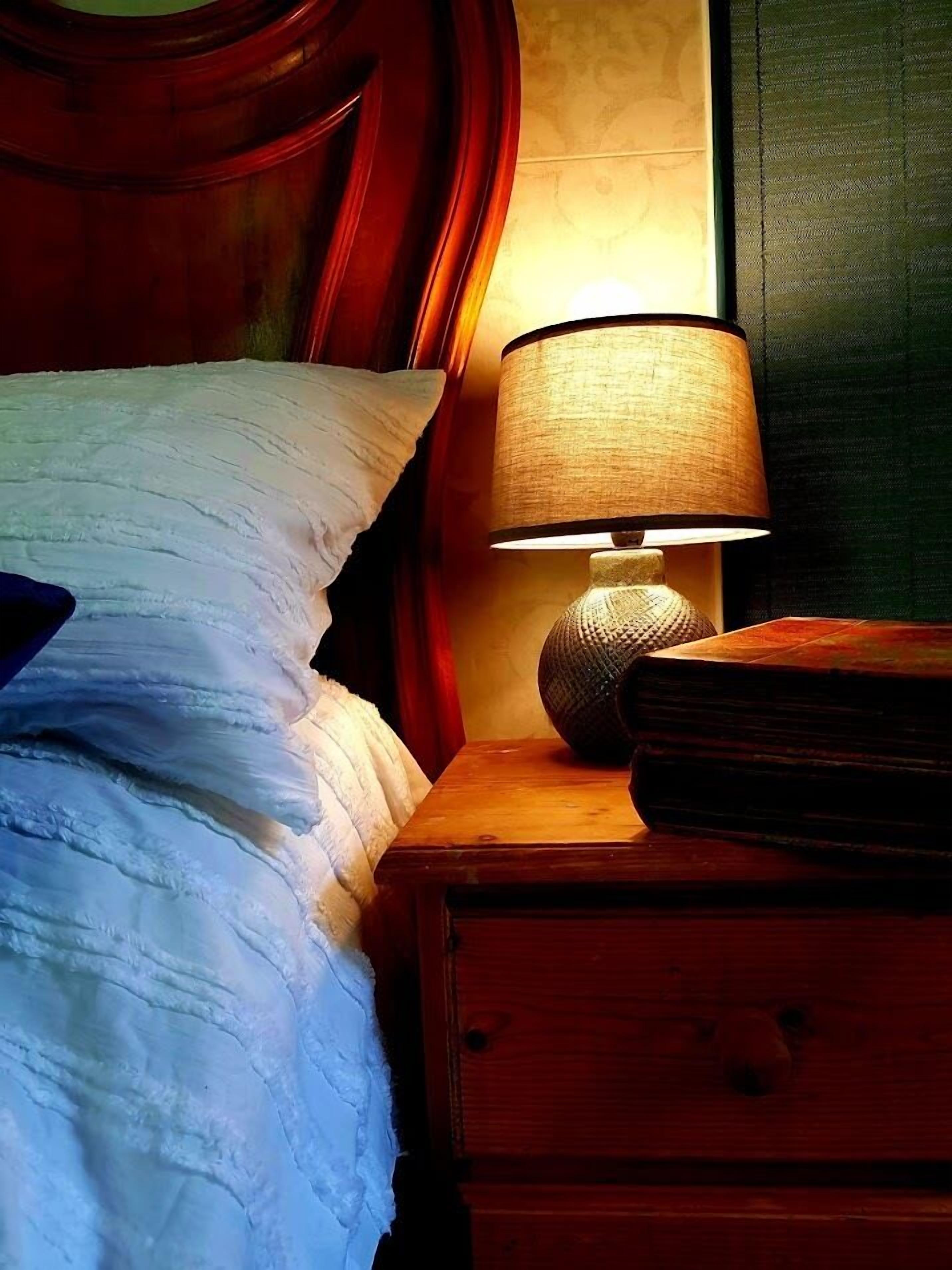 A bed with a textured white duvet, a wooden nightstand with a lamp, and a book.
