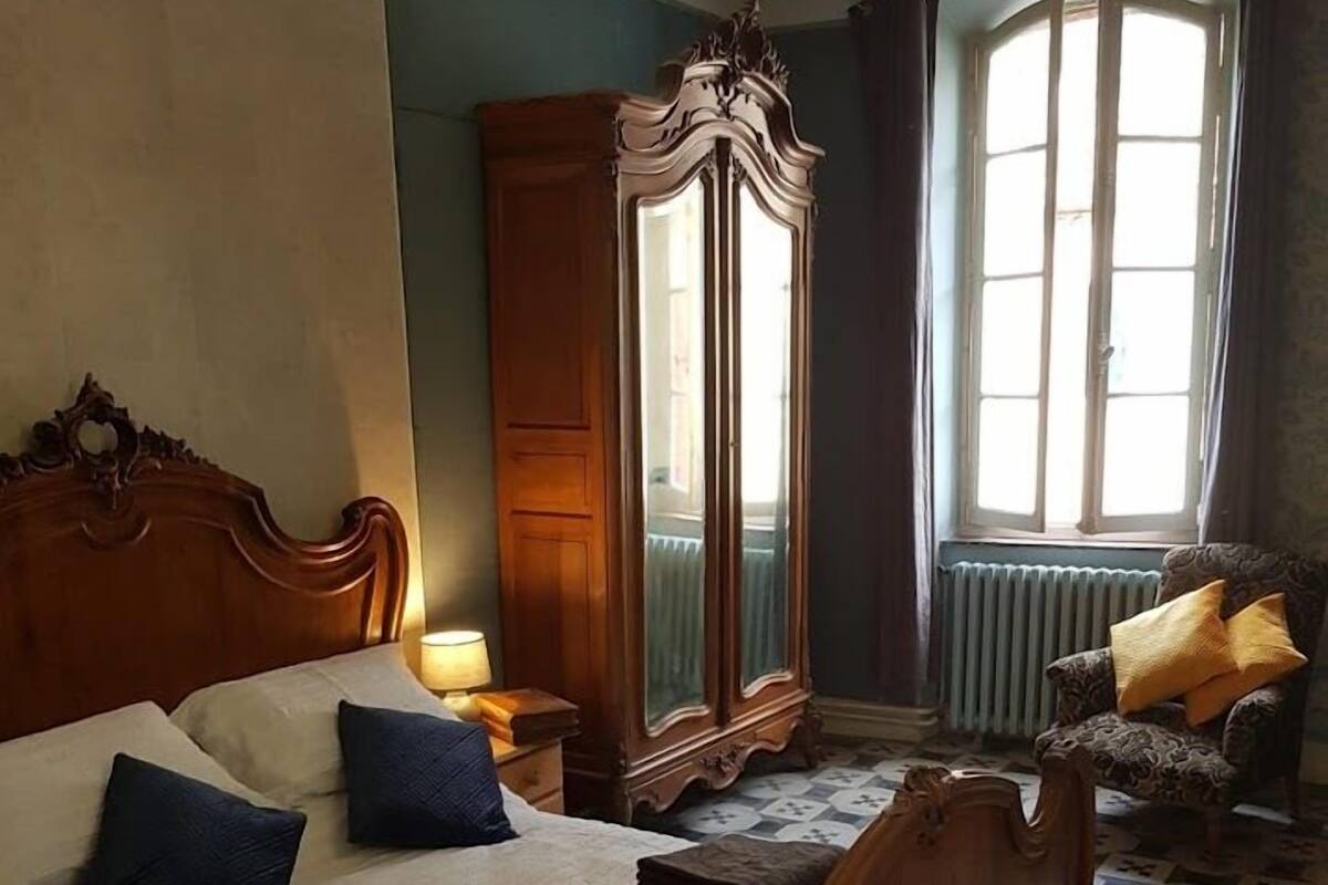 A bedroom with a large bed, a wooden wardrobe, a mirror, a chair, and a window with curtains.