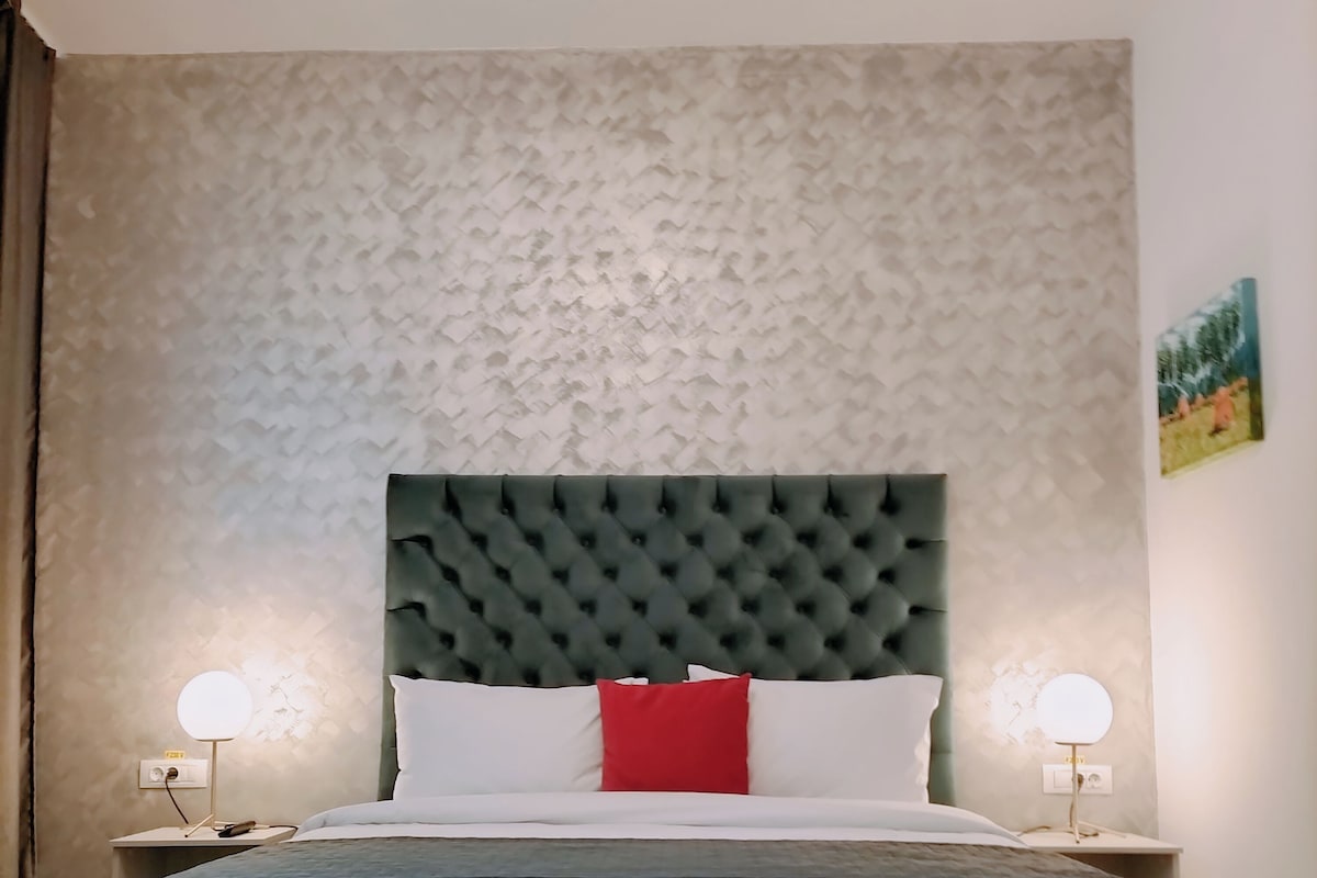 A neatly arranged bedroom with a tufted headboard, a bed with white and red bedding, two bedside lamps, and a patterned wall.