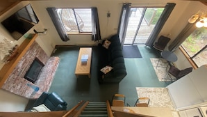 A spacious living area with a brick fireplace, a sofa, a coffee table, and a TV mounted on the wall. There are two windows with curtains, a staircase leading to another level, and a small table with chairs. - Waterfront cozy cabin vacation home with dock (Bamfield)